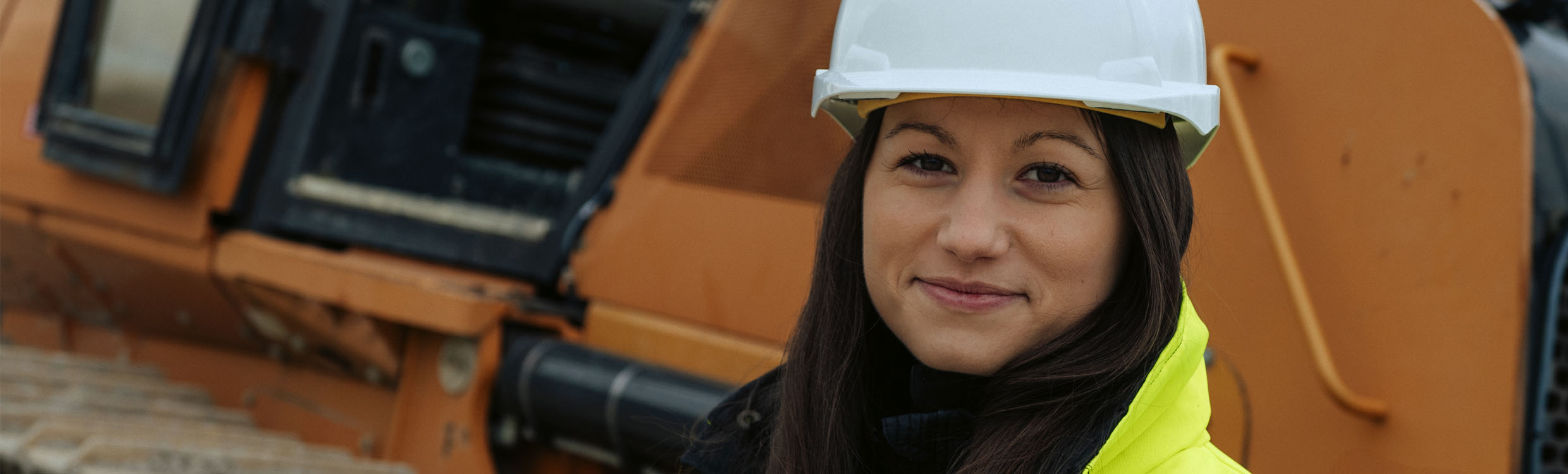 Female construction worker with white helmet and safety vest in front of a dozer. 